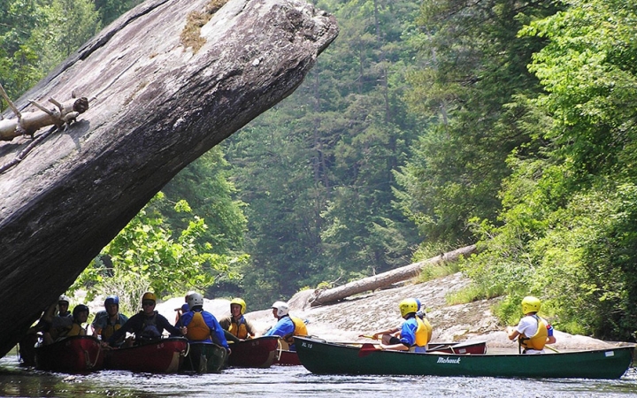 People sit in canoes resting in the shade of a large rock.
